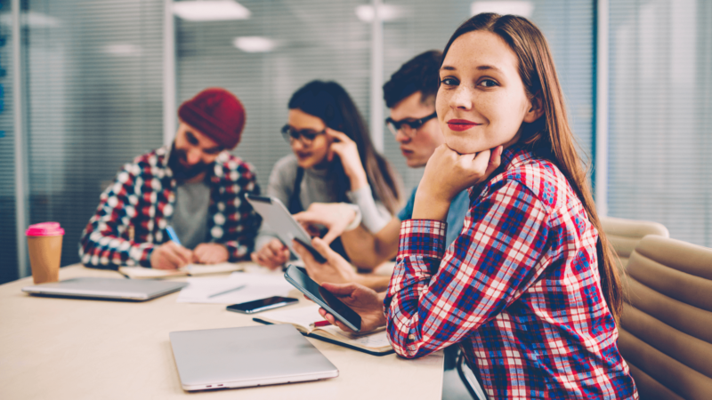 A woman with long brown hair in a red plaid shirt sitting confidently in a group setting after attending a Christian Counseling & Coaching session, smiling at the camera while her peers work in the background.