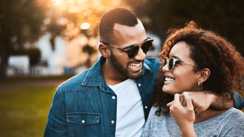 A couple smiling while walking together outdoors, both wearing sunglasses and enjoying a sunny day.