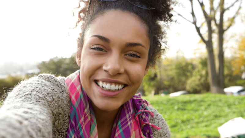 Smiling woman outdoors, wearing a colorful scarf, enjoying a sunny day in a park.