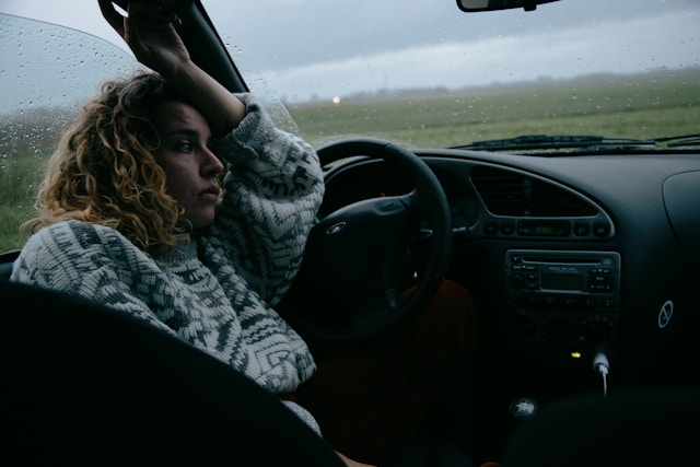 A thoughtful woman sitting in a parked car on a rainy day, gazing out the window, symbolizing introspection and overthinking.